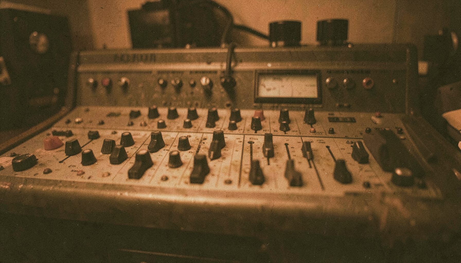 Warm-toned analog film photograph of a vintage mixing console with worn faders and knobs, bathed in low amber light, visible film grain and soft shadows across the panel.