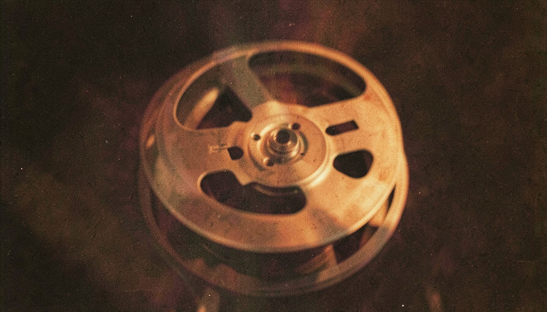 Close-up of a vintage film reel against a dark background, soft golden highlights on the metal spool, grainy analog texture