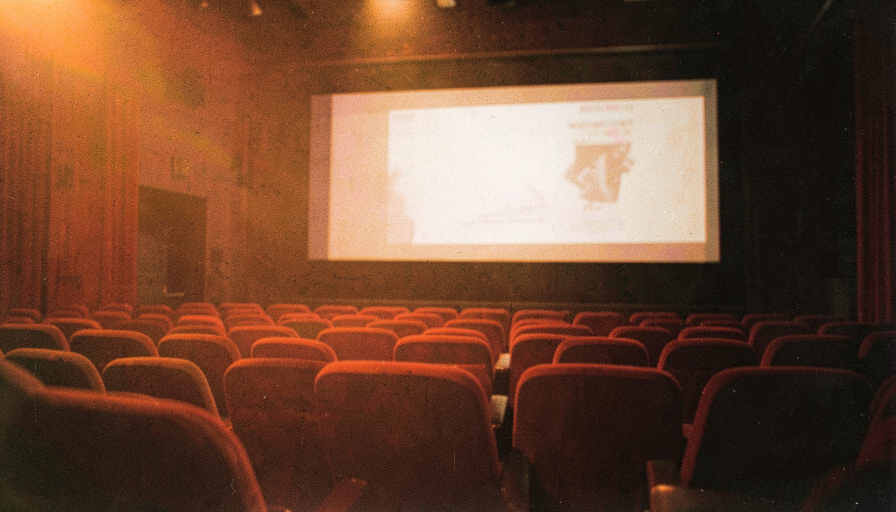Dim theater interior with deep red velvet seats and a glowing projection screen, warm amber light spilling across the room, grainy film-stock texture