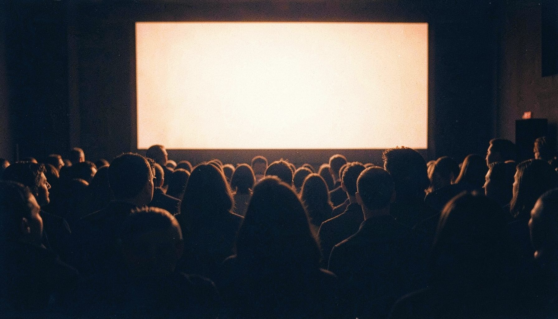 Crowded cinema interior seen from behind, heads silhouetted against a bright screen, warm amber and deep blue tones, grainy film aesthetic