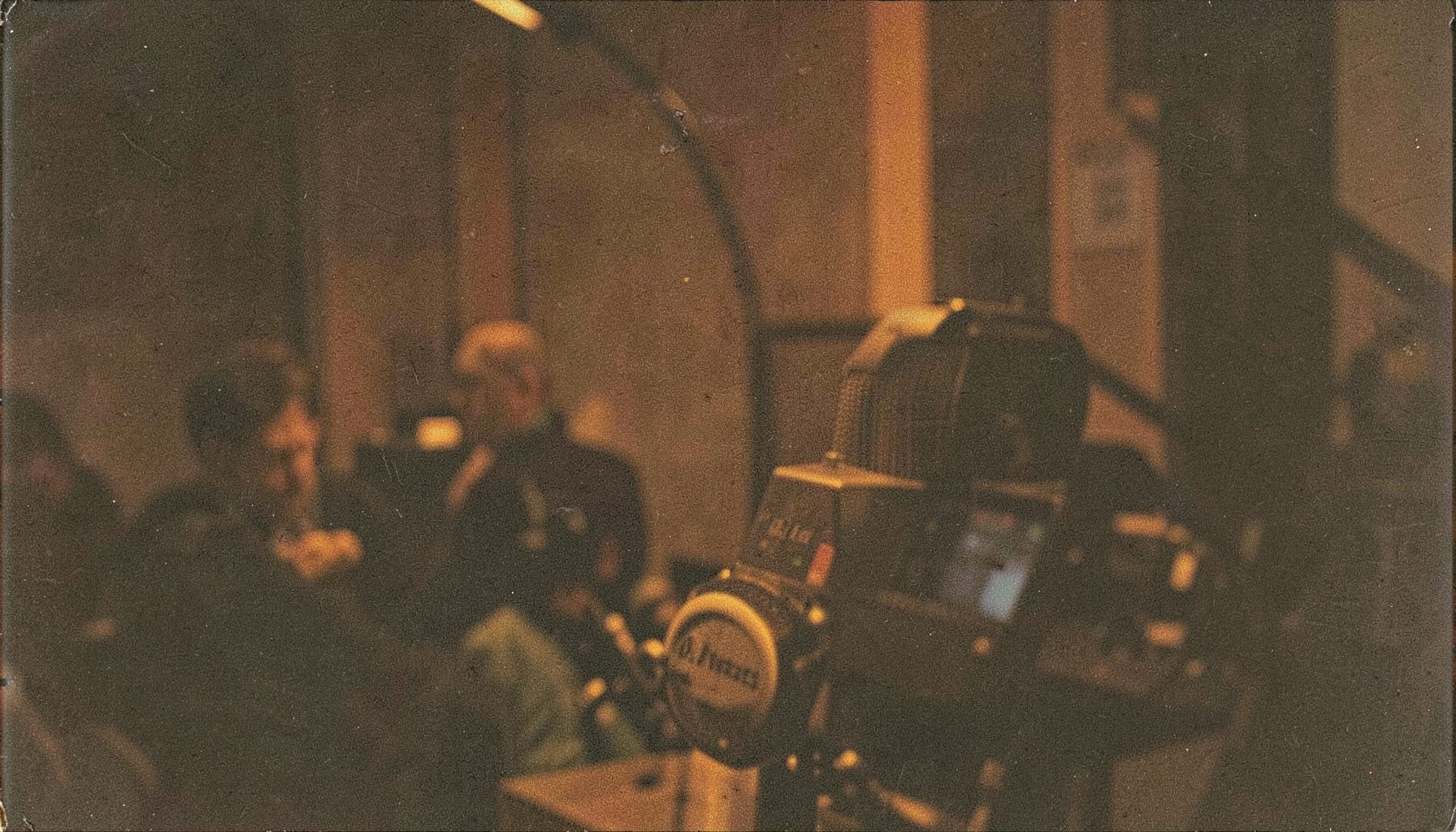 A grainy analog photograph of a dimly lit radio studio with a microphone in the foreground, warm amber and deep brown tones, moody and intimate atmosphere.