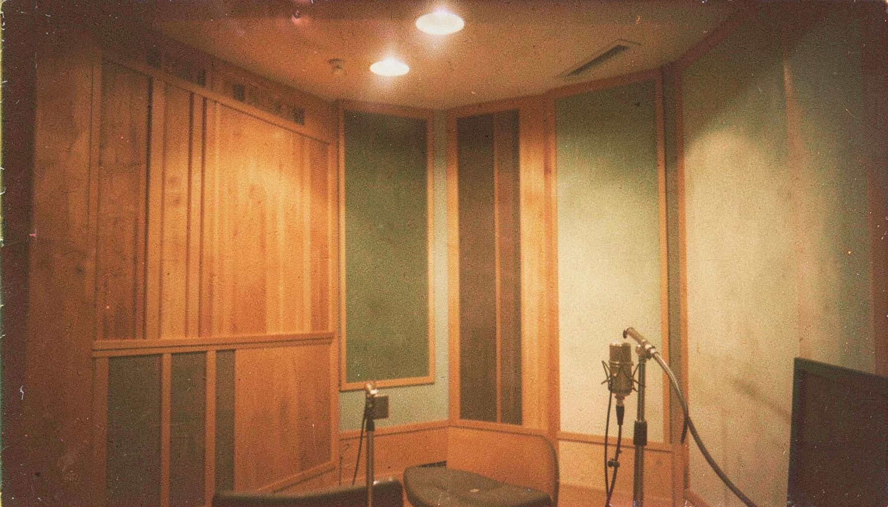 A textured film photo of a recording studio interior with warm wood panels and soft overhead lighting, a microphone stand in the foreground against muted green and cream walls.