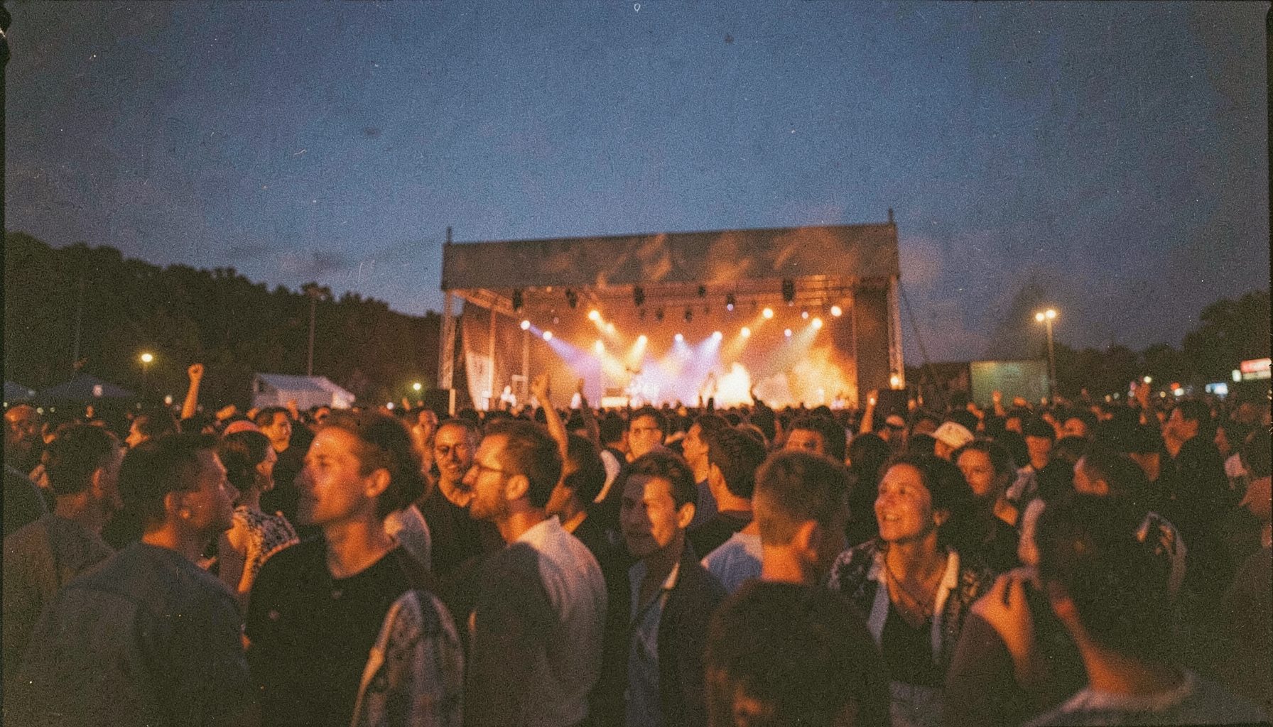 A grainy analog photograph of an outdoor music festival crowd at dusk, with deep indigo skies and warm orange stage lights reflecting off faces, conveying a communal celebration.