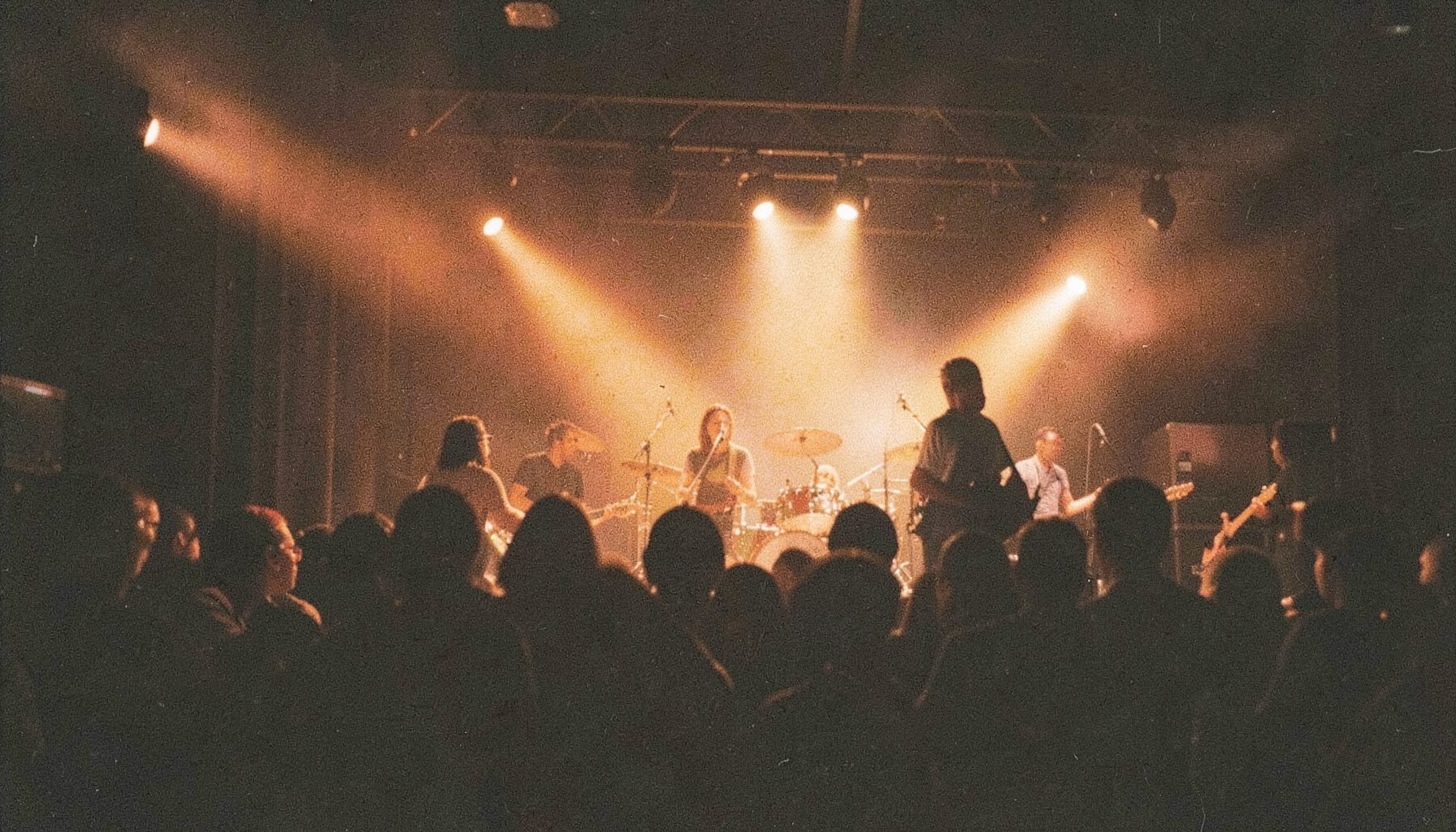 A grainy film photograph of a live band on a warmly lit stage, with deep brown and gold tones, musicians silhouetted against amber spotlights, capturing the energy of an East African concert.
