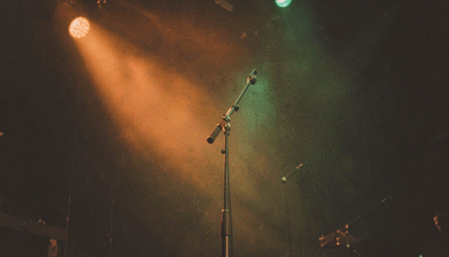 A warm, grainy film photograph of a dimly lit concert stage with amber and deep green stage lights casting soft glows across an empty microphone stand, capturing the quiet anticipation before a live East African music performance.
