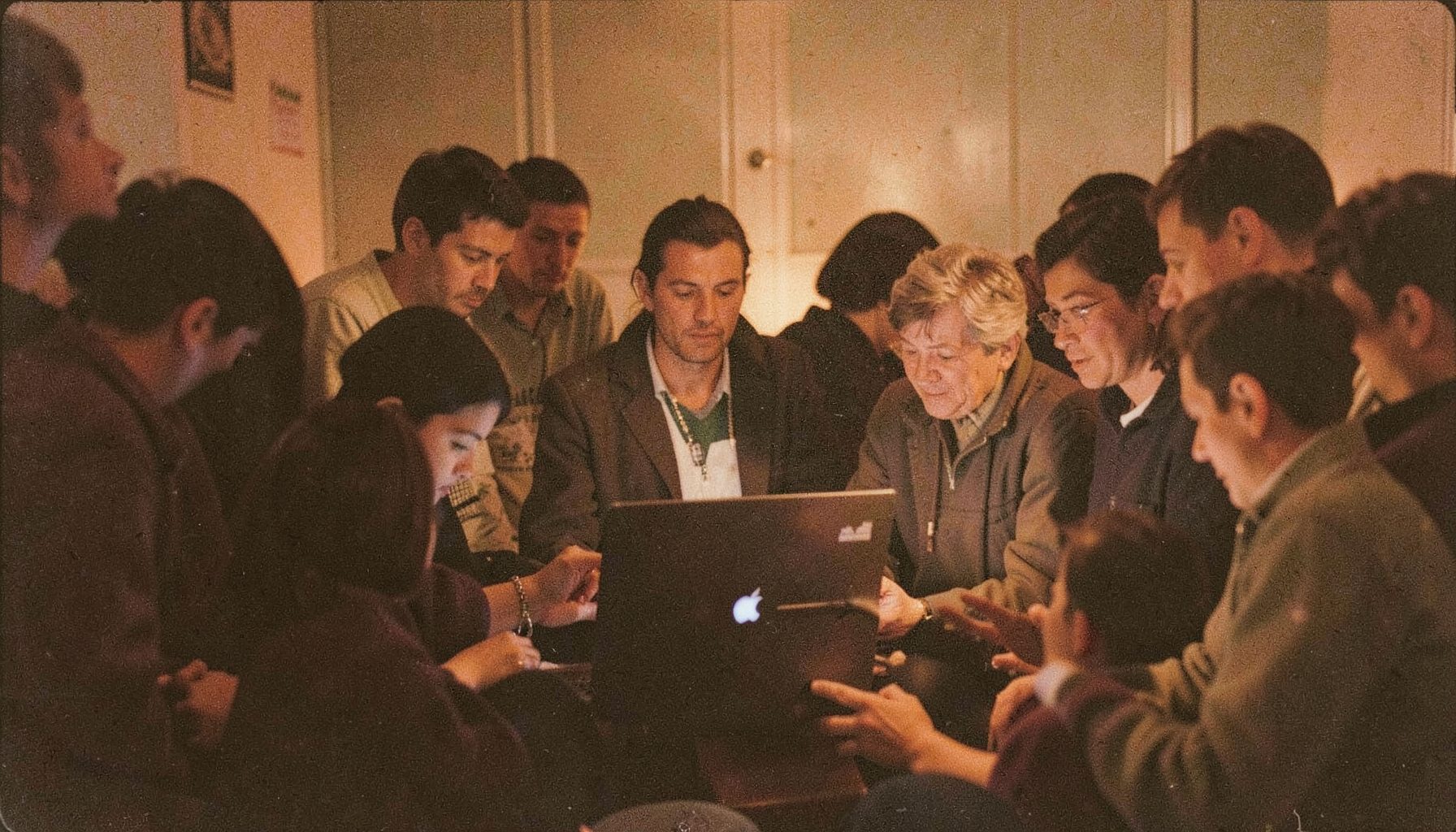 A warm, grainy film photograph of a small crowd gathered around a laptop in a softly lit room, deep browns and muted gold tones, intimate community atmosphere