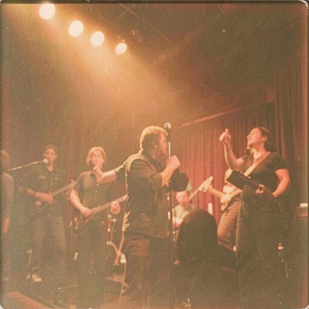 Grainy analog film photograph of a crowded stage performance with warm brown stage lighting and silhouetted figures